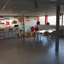 Deze lichte en gezellige kantine heeft twee grote tafels met houten stoelen en een bar met hoge krukken. Kleurrijke borden en decoraties, zoals luchtballonfiguurtjes en een rode kroonluchter, voegen een speels accent toe. Grote ramen zorgen voor veel licht, terwijl de eenvoudige inrichting ideaal is voor ontspanning, bijeenkomsten of activiteiten.