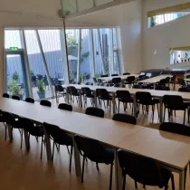 Een grote, moderne zaal met lange tafels en zwarte stoelen, ideaal voor bijeenkomsten of evenementen. De ruimte heeft hoge ramen die veel natuurlijk licht binnenlaten en uitzicht bieden op een groen buitenterras. Planten en kleurrijke decoraties langs de ramen voegen een frisse, uitnodigende sfeer toe. De lichte vloer en muren versterken het open en ruimtelijke karakter.