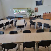 Een ruime, multifunctionele zaal met meerdere opstellingen van tafels en stoelen, ideaal voor groepsbijeenkomsten of evenementen. De ruimte is licht dankzij grote ramen en heeft een warme uitstraling door houten elementen, zoals een piano en een gele boekenkast. Planten en decoratieve rode potten voegen een frisse, uitnodigende sfeer toe, terwijl de tv en de lampen het geheel functioneel en modern maken.