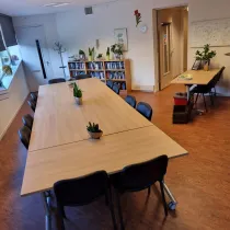 Een sfeervolle vergaderruimte met een lange houten tafel, omringd door comfortabele zwarte stoelen. Planten en kleurrijke raamdecoraties voegen een frisse en levendige touch toe. Aan de zijkant staan boekenplanken gevuld met boeken, en er is een extra bureau met stoelen voor individueel werk. De warme houten vloer en praktische inrichting creëren een uitnodigende, functionele sfeer voor zowel vergaderingen als creatieve sessies.