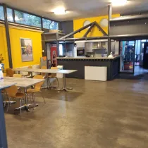 Een industriële kantine met een gele muur, open keuken en verspreide tafels. Grote ramen zorgen voor licht, terwijl de inrichting praktisch en functioneel oogt.