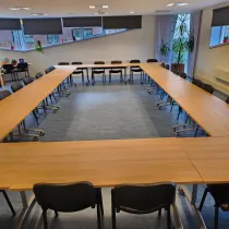 Een ruime vergaderruimte met een grote U-vormige tafelopstelling, omringd door zwarte stoelen. De kamer is licht en modern, met grote ramen die zorgen voor veel natuurlijk licht. Planten en decoratieve elementen voegen een frisse en uitnodigende sfeer toe. De ruimte is ideaal voor grote bijeenkomsten, presentaties of workshops.