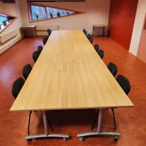 Een nette vergaderruimte met een lange houten tafel, omringd door zwarte stoelen. De schuine ramen met planten op de vensterbanken zorgen voor natuurlijk licht en een frisse uitstraling. De warme, rode vloer en opvallende rode kastwand geven de ruimte een levendige en functionele sfeer, ideaal voor formele bijeenkomsten of teamoverleg.