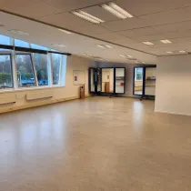 Een ruime, open zaal met grote ramen die veel natuurlijk licht binnenlaten en uitzicht bieden op een buitenomgeving. De lichte vloer en het strakke, witte plafond creëren een frisse en veelzijdige ruimte, ideaal voor activiteiten zoals vergaderingen, workshops of bewegingslessen. Verplaatsbare spiegels aan de achterkant voegen flexibiliteit toe aan de inrichting, terwijl de moderne uitstraling de ruimte functioneel en uitnodigend maakt.