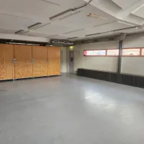 Een ruime multifunctionele zaal met een strak en eenvoudig ontwerp. Grote houten opbergkasten bieden praktische opslagmogelijkheden, terwijl de lange ramen voor natuurlijk licht zorgen. De open indeling maakt de ruimte flexibel en geschikt voor verschillende activiteiten zoals workshops, bijeenkomsten of sportlessen.