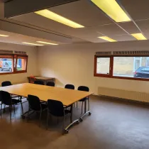 Een ruime vergaderruimte met een grote houten tafel, omringd door blauwe stoelen. Grote ramen zorgen voor natuurlijk licht en een open sfeer, terwijl ingebouwde verlichting de ruimte helder en functioneel maakt.
