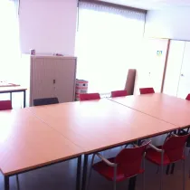 Een lichte en functionele vergaderruimte met een grote, rechthoekige tafel omringd door rode en zwarte stoelen. Grote ramen met witte gordijnen zorgen voor veel natuurlijk licht en een open sfeer. Aan de zijkant staat een opbergkast en extra tafels, wat de ruimte praktisch en veelzijdig maakt. De minimalistische inrichting maakt het ideaal voor vergaderingen, workshops of kleine bijeenkomsten.