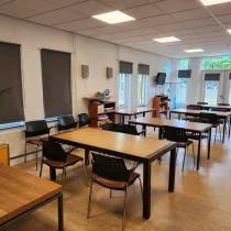 De huiskamer in het Buitenhuis is een lichte en gezellige ruimte met houten tafels, comfortabele stoelen en grote ramen. Voorzien van een televisie, boekenplank en directe toegang tot buiten, ideaal voor diverse activiteiten en bijeenkomsten.