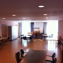Een veelzijdige en ruime zaal met verschillende tafels en stoelen, ideaal voor groepsactiviteiten of bijeenkomsten. Grote ramen met gordijnen laten veel natuurlijk licht binnen, wat de ruimte open en uitnodigend maakt. Een rij kluisjes staat langs de muur voor praktische opslag, terwijl extra stoelen en een kapstok achterin de zaal flexibiliteit bieden. De warme tinten van de vloer en het meubilair creëren een comfortabele en functionele sfeer.
