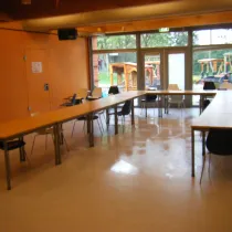 Een ruime zaal met lange tafels in een flexibele opstelling, ideaal voor vergaderingen of workshops. Grote ramen en glazen deuren bieden veel lichtinval en zicht op een buitenspeelplaats. De warme muurkleuren en houten accenten creëren een aangename, uitnodigende sfeer.