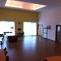 Een ruime en sfeervolle zaal met een hoge gele accentmuur en grote ramen voorzien van lichte gordijnen, die veel natuurlijk licht binnenlaten. In het midden staat een biljarttafel, wat de ruimte een sociale en multifunctionele functie geeft. Hangende lampen voegen een warm accent toe, terwijl tafels, stoelen en een verrijdbare kapstok aan de zijkant de zaal flexibel en praktisch in gebruik maken. Ideaal voor bijeenkomsten, ontspanning of evenementen.