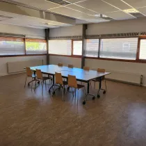 Een lichte en open activiteitenruimte met grote ramen die natuurlijk licht binnenlaten. De ruimte is voorzien van een centrale tafel met stoelen, geschikt voor vergaderingen, workshops of kleine groepsactiviteiten. Het eenvoudige interieur en de flexibele indeling maken het een veelzijdige plek voor diverse doeleinden.