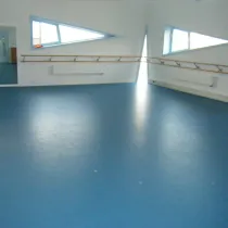 Een lichte en ruime oefenruimte met een blauwe vloer en strakke witte muren. De grote spiegels aan de ene kant en de houten barre langs de muren maken de zaal perfect voor dans- of bewegingslessen. De driehoekige ramen zorgen voor natuurlijk licht en geven de ruimte een moderne uitstraling.