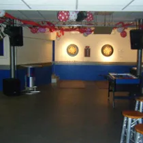  Deze jongerenruimte heeft een moderne, feestelijke uitstraling met blauwe muren en rode en roze versieringen aan het plafond. In het midden hangen twee verlichte dartborden met een scorebord ertussen, perfect voor een potje darten. Links staat een Red Bull-koeler, terwijl rechts een voetbaltafel en hoge krukken rond een bar zichtbaar zijn. Grote speakers en spotlampen wijzen op mogelijkheden voor muziek en feestjes. De ruimte straalt gezelligheid uit en biedt plek voor ontspanning, spel en samenzijn.