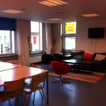 Een gezellige huiskamerruimte met een uitnodigende zithoek bestaande uit een bank en kleurrijke kussens, een laag salontafeltje en comfortabele fauteuils. De ruimte heeft een warme sfeer dankzij de houten tafels, stoelen en het kleurrijke tapijt. Grote ramen met gordijnen zorgen voor veel natuurlijk licht, terwijl een muur met een tv en een krijtbord de functionaliteit versterken. Deze ruimte is ideaal voor informele bijeenkomsten, ontspanning of sociale activiteiten.