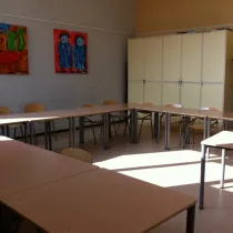Een lichte en ruime activiteitenruimte met een U-vormige tafelopstelling en houten stoelen eromheen, ideaal voor vergaderingen of groepsactiviteiten. De hoge kasten aan de achterkant bieden praktische opbergruimte, terwijl kleurrijke schilderijen aan de muur de ruimte een creatieve en levendige uitstraling geven. De grote ramen zorgen voor veel natuurlijk licht, wat de ruimte uitnodigend en prettig maakt.