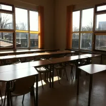 Een lichte les- of activiteitenruimte met grote ramen die een panoramisch uitzicht bieden en zorgen voor een overvloed aan natuurlijk licht. De tafels en stoelen zijn georganiseerd in een flexibele opstelling, ideaal voor onderwijs of groepswerk. De zachte oranje gordijnen voegen een warme en uitnodigende sfeer toe. De ruimte heeft een praktische, functionele uitstraling, geschikt voor diverse activiteiten.