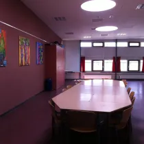 Een stijlvolle zaal met een warme, donkerrode accentmuur, voorzien van kleurrijke kunstwerken die de ruimte een creatieve uitstraling geven. Grote ramen met rode gordijnen zorgen voor natuurlijk licht en bieden een warme sfeer. Een lange tafel met houten stoelen in het midden maakt de ruimte ideaal voor vergaderingen of groepsactiviteiten. De plafondverlichting, draagt bij aan een moderne en goed verlichte omgeving, terwijl de hoge kast aan de zijkant zorgt voor praktische opbergruimte.