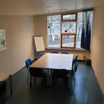 Een compacte en functionele vergaderruimte met een eenvoudige opstelling van tafels en blauwe stoelen, geschikt voor kleine groepen. Het grote raam laat veel natuurlijk licht binnen en biedt uitzicht op een rustige straat. Aan de muur hangt een kunstwerk dat kleur en persoonlijkheid toevoegt aan de ruimte. Een flip-over in de hoek benadrukt de praktische bruikbaarheid van de ruimte voor brainstormsessies of presentaties. De kamer heeft een rustige, professionele uitstraling.