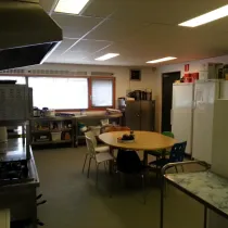 Een volledig uitgeruste keuken met een praktische indeling. Voorzien van een groot fornuis, opslagrekken en meerdere koelkasten. In het midden staat een ronde tafel met stoelen, ideaal voor informele bijeenkomsten of voorbereiding. De ruimte is functioneel en goed verlicht, geschikt voor kookactiviteiten of catering.