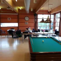 Een gezellige ontmoetingsruimte met een biljarttafel in het midden, omgeven door tafels en stoelen. Grote ramen laten veel licht binnen en bieden uitzicht op het groen. Warme houten accenten en hanglampen zorgen voor een sfeervolle uitstraling.