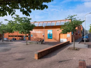 Het gebouw heeft een robuuste, hoekige uitstraling met warme bakstenen gevel en horizontale lijnen, ontworpen met een verfijnd contrast tussen stenen buitengevels en subtiel naar voren stekende houten volumes. Met een luifel boven de ingang en grote glaspartijen oogt het gebouw uitnodigend en afgestemd op het omliggende groene plein. 