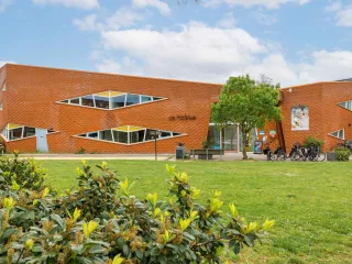 Een opvallend, modern wijkgebouw met rode baksteenstructuur en speelse driehoekige ramen. De schuine lijnen en asymmetrische vormen geven het gebouw een dynamische uitstraling. Voor het gebouw ligt een ruim plein met enkele plantenbakken, banken en een geparkeerde fiets, wat een functionele en toegankelijke sfeer creëert.