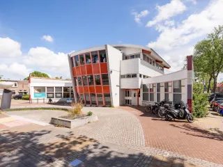 Een opvallend jaren ’70/’80-gebouw met een strakke, functionele architectuur bestaande uit witte gevels met grote raamstroken en enkele kleuraccenten in rood, zichtbaar in zowel de enkele verdieping als de deels twee- of driekleurige bouwdelen
