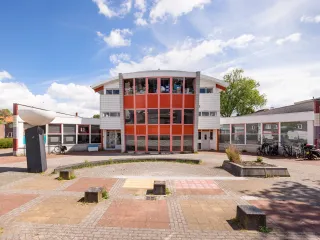 Een opvallend jaren ’70/’80-gebouw met een strakke, functionele architectuur bestaande uit witte gevels met grote raamstroken en enkele kleuraccenten in rood, zichtbaar in zowel de enkele verdieping als de deels twee- of driekleurige bouwdelen