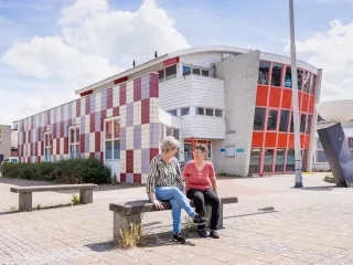 Een opvallend jaren ’70/’80-gebouw met een strakke, functionele architectuur bestaande uit witte gevels met grote raamstroken en enkele kleuraccenten in rood, zichtbaar in zowel de enkele verdieping als de deels twee- of driekleurige bouwdelen