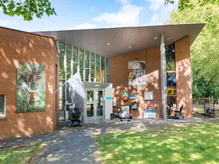 Een modern gebouw met een bakstenen gevel en grote glaspartij bij de entree, wat zorgt voor een open en uitnodigende uitstraling. Aan de voorzijde loopt een strak betegeld pad langs een groene grasstrook en grote bomen die voor schaduw zorgen. Een mooi entreebord naast de ingang duidt op activiteiten binnen het gebouw. De combinatie van natuur en architectuur creëert een rustige, aangename sfeer.