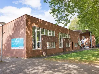 Een modern gebouw met een bakstenen gevel en grote glaspartij bij de entree, wat zorgt voor een open en uitnodigende uitstraling. Aan de voorzijde loopt een strak betegeld pad langs een groene grasstrook en grote bomen die voor schaduw zorgen. Een mooi entreebord naast de ingang duidt op activiteiten binnen het gebouw. De combinatie van natuur en architectuur creëert een rustige, aangename sfeer.