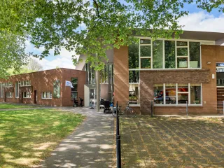 Een modern gebouw met een bakstenen gevel en grote glaspartij bij de entree, wat zorgt voor een open en uitnodigende uitstraling. Aan de voorzijde loopt een strak betegeld pad langs een groene grasstrook en grote bomen die voor schaduw zorgen. Een mooi entreebord naast de ingang duidt op activiteiten binnen het gebouw. De combinatie van natuur en architectuur creëert een rustige, aangename sfeer.