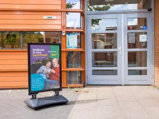 Het gebouw heeft een robuuste, hoekige uitstraling met warme bakstenen gevel en horizontale lijnen, ontworpen met een verfijnd contrast tussen stenen buitengevels en subtiel naar voren stekende houten volumes. Met een luifel boven de ingang en grote glaspartijen oogt het gebouw uitnodigend en afgestemd op het omliggende groene plein. 