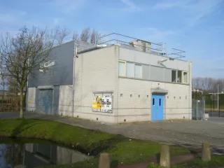 Een modern, rechthoekig gebouw met witte bakstenen muren en een blauwe toegangsdeur. De bovenverdieping heeft meerdere ramen en een metalen installatie op het dak. Voor het gebouw ligt een betegeld plein, met aan de zijkant een grasstrook en een sloot die de weerspiegeling van het gebouw laat zien. Een kale boom staat links van het gebouw, terwijl de lucht helder en blauw is.