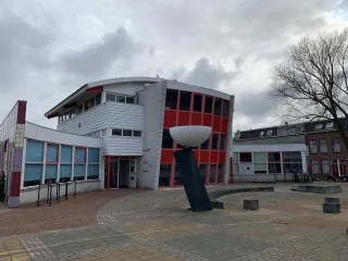 Een opvallend jaren ’70/’80-gebouw met een strakke, functionele architectuur bestaande uit witte gevels met grote raamstroken en enkele kleuraccenten in rood, zichtbaar in zowel de enkele verdieping als de deels twee- of driekleurige bouwdelen
