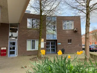 De Wippolder in Delft is een eigentijds ogend, laagbouwpand met een robuuste bakstenen gevel, waarbij de entree subtiel is benadrukt met een overstekend dakje en grote glaspuien die uitnodigend uitzicht bieden op de omliggende groenrijke pleinruimte.