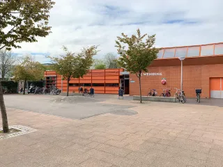 Het gebouw heeft een robuuste, hoekige uitstraling met warme bakstenen gevels en horizontale lijnen, ontworpen met een verfijnd contrast tussen stenen buitengevels en subtiel naar voren stekende houten volumes. Met een luifel boven de ingang en grote glaspartijen oogt het gebouw uitnodigend en afgestemd op het omliggende groene plein.