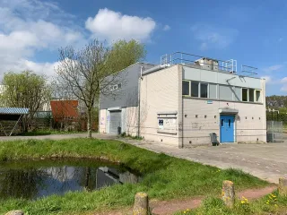 Een modern, rechthoekig gebouw met witte bakstenen muren en een blauwe toegangsdeur. De bovenverdieping heeft meerdere ramen en een metalen installatie op het dak. Voor het gebouw ligt een betegeld plein, met aan de zijkant een grasstrook en een sloot die de weerspiegeling van het gebouw laat zien. Een kale boom staat links van het gebouw, terwijl de lucht helder en blauw is.
