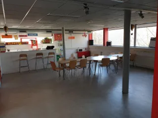 Deze lichte en gezellige kantine heeft twee grote tafels met houten stoelen en een bar met hoge krukken. Kleurrijke borden en decoraties, zoals luchtballonfiguurtjes en een rode kroonluchter, voegen een speels accent toe. Grote ramen zorgen voor veel licht, terwijl de eenvoudige inrichting ideaal is voor ontspanning, bijeenkomsten of activiteiten.
