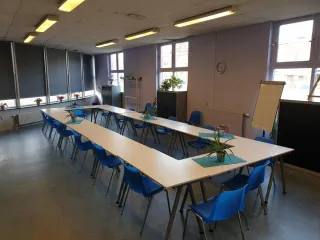 Een creatieve en multifunctionele ruimte met een open en lichte sfeer. De lange tafels in een U-vorm opstelling bieden plaats aan groepsactiviteiten zoals workshops, vergaderingen of cursussen. De blauwe stoelen geven een kleurrijke touch aan de ruimte. Aan de achterkant bevindt zich een kitchenette, handig voor het bereiden van koffie of thee. De kunstwerken en schildersezels langs de muur wijzen op een creatieve omgeving, ideaal voor kunst- en handvaardigheidssessies.