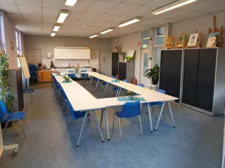 Een creatieve en multifunctionele ruimte met een open en lichte sfeer. De lange tafels in een U-vorm opstelling bieden plaats aan groepsactiviteiten zoals workshops, vergaderingen of cursussen. De blauwe stoelen geven een kleurrijke touch aan de ruimte. Aan de achterkant bevindt zich een kitchenette, handig voor het bereiden van koffie of thee. De kunstwerken en schildersezels langs de muur wijzen op een creatieve omgeving, ideaal voor kunst- en handvaardigheidssessies.
