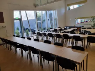 Een grote, moderne zaal met lange tafels en zwarte stoelen, ideaal voor bijeenkomsten of evenementen. De ruimte heeft hoge ramen die veel natuurlijk licht binnenlaten en uitzicht bieden op een groen buitenterras. Planten en kleurrijke decoraties langs de ramen voegen een frisse, uitnodigende sfeer toe. De lichte vloer en muren versterken het open en ruimtelijke karakter.