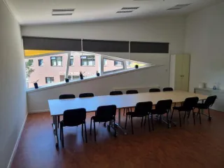 Een moderne vergaderruimte met een grote tafel en meerdere stoelen, perfect voor groepsbijeenkomsten. Het opvallende driehoekige raam laat veel natuurlijk licht binnen en biedt uitzicht op een aangrenzend gebouw. De minimalistische inrichting met enkele planten op de vensterbank geeft de ruimte een frisse, professionele uitstraling.