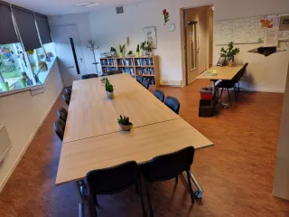 Een sfeervolle vergaderruimte met een lange houten tafel, omringd door comfortabele zwarte stoelen. Planten en kleurrijke raamdecoraties voegen een frisse en levendige touch toe. Aan de zijkant staan boekenplanken gevuld met boeken, en er is een extra bureau met stoelen voor individueel werk. De warme houten vloer en praktische inrichting creëren een uitnodigende, functionele sfeer voor zowel vergaderingen als creatieve sessies.