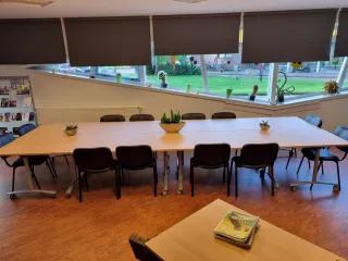 Een sfeervolle vergaderruimte met een lange houten tafel, omringd door comfortabele zwarte stoelen. Planten en kleurrijke raamdecoraties voegen een frisse en levendige touch toe. Aan de zijkant staan boekenplanken gevuld met boeken, en er is een extra bureau met stoelen voor individueel werk. De warme houten vloer en praktische inrichting creëren een uitnodigende, functionele sfeer voor zowel vergaderingen als creatieve sessies.