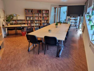 Een sfeervolle vergaderruimte met een lange houten tafel, omringd door comfortabele zwarte stoelen. Planten en kleurrijke raamdecoraties voegen een frisse en levendige touch toe. Aan de zijkant staan boekenplanken gevuld met boeken, en er is een extra bureau met stoelen voor individueel werk. De warme houten vloer en praktische inrichting creëren een uitnodigende, functionele sfeer voor zowel vergaderingen als creatieve sessies.