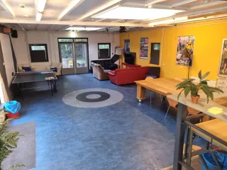 Een multifunctionele ruimte met een speelse, blauwe vloer en een opvallende gele muur. De inrichting bestaat uit tafels, stoelen, een tafeltennistafel en een zithoek met banken, wat een ontspannen en sociale sfeer creëert. Grote ramen en deuren zorgen voor veel natuurlijk licht.