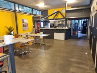 Een industriële kantine met een gele muur, open keuken en verspreide tafels. Grote ramen zorgen voor licht, terwijl de inrichting praktisch en functioneel oogt.