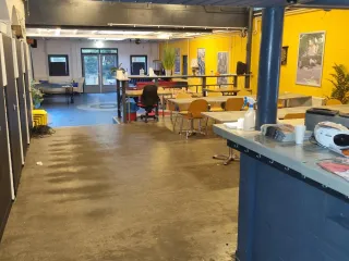 Een ruime, industriële kantine met een gele accentmuur, tafels en stoelen verspreid over de ruimte. De betonnen vloer, open keuken en grote ramen geven de ruimte een functionele en moderne uitstraling.